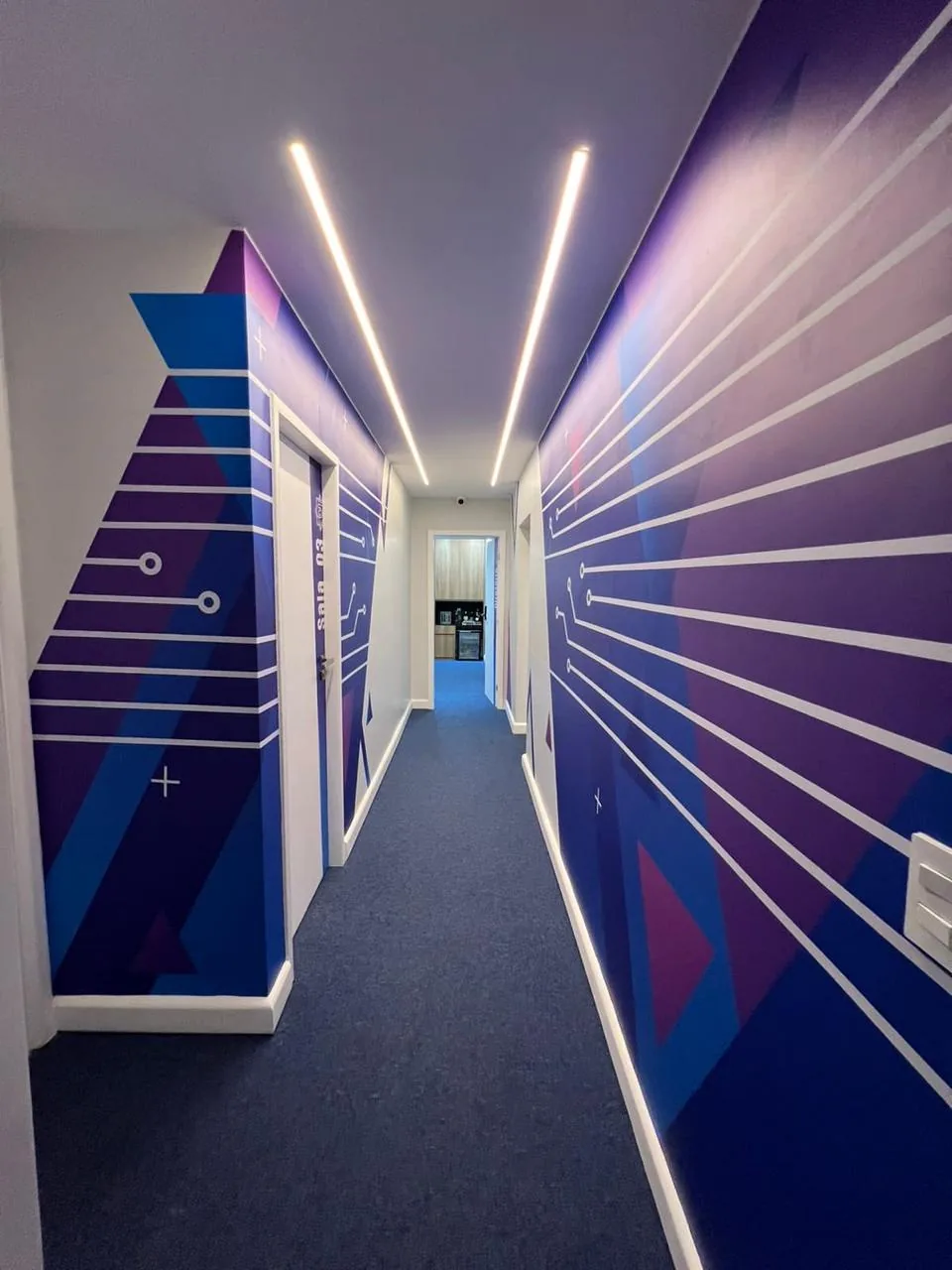 Corredor com paredes personalizadas em azul e roxo e iluminação linear no teto.